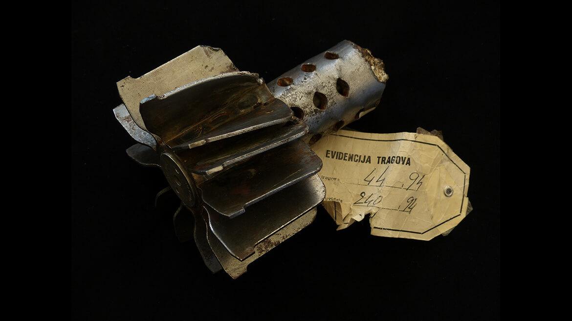 Teil einer Mörsergranate als Beweisstück vor dem Internationalen Strafgerichtshof zu Jugoslawien, Den Haag: Im Bosnischen Bürgerkrieg belagern Regierungstruppen die Stadt Sarajevo fast vier Jahre lang. Diese Granate wird am 05.02.1994 von Regierungstruppen auf die Markthalle in Sarajevo abgefeuert und tötet 60 Zivilistinnen und Zivilisten.