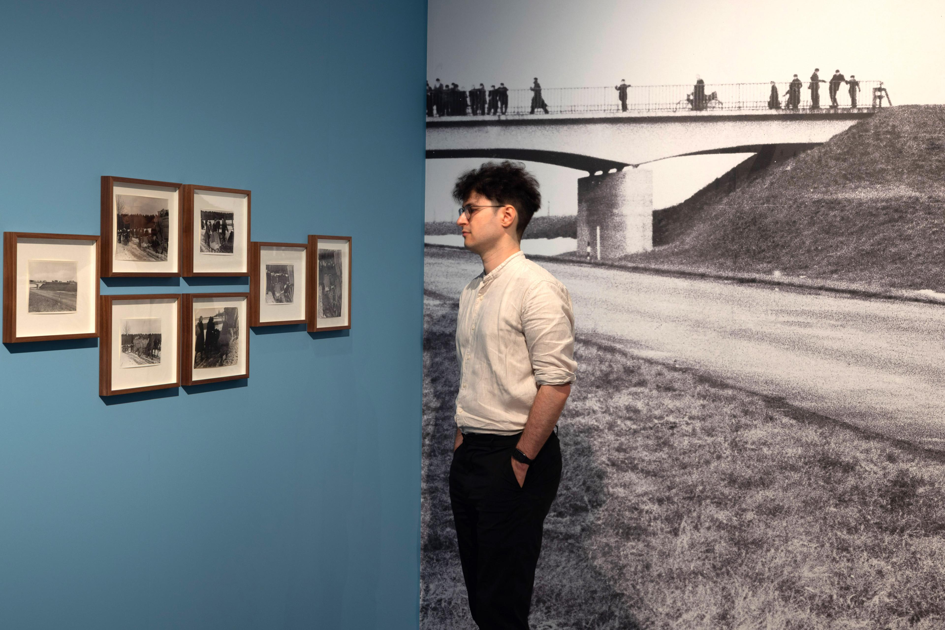 Ausstellungsansicht von "Der Treck". Ein Mann steht vor einer blauen Wand, an der eingerahmte Fotos hängen.