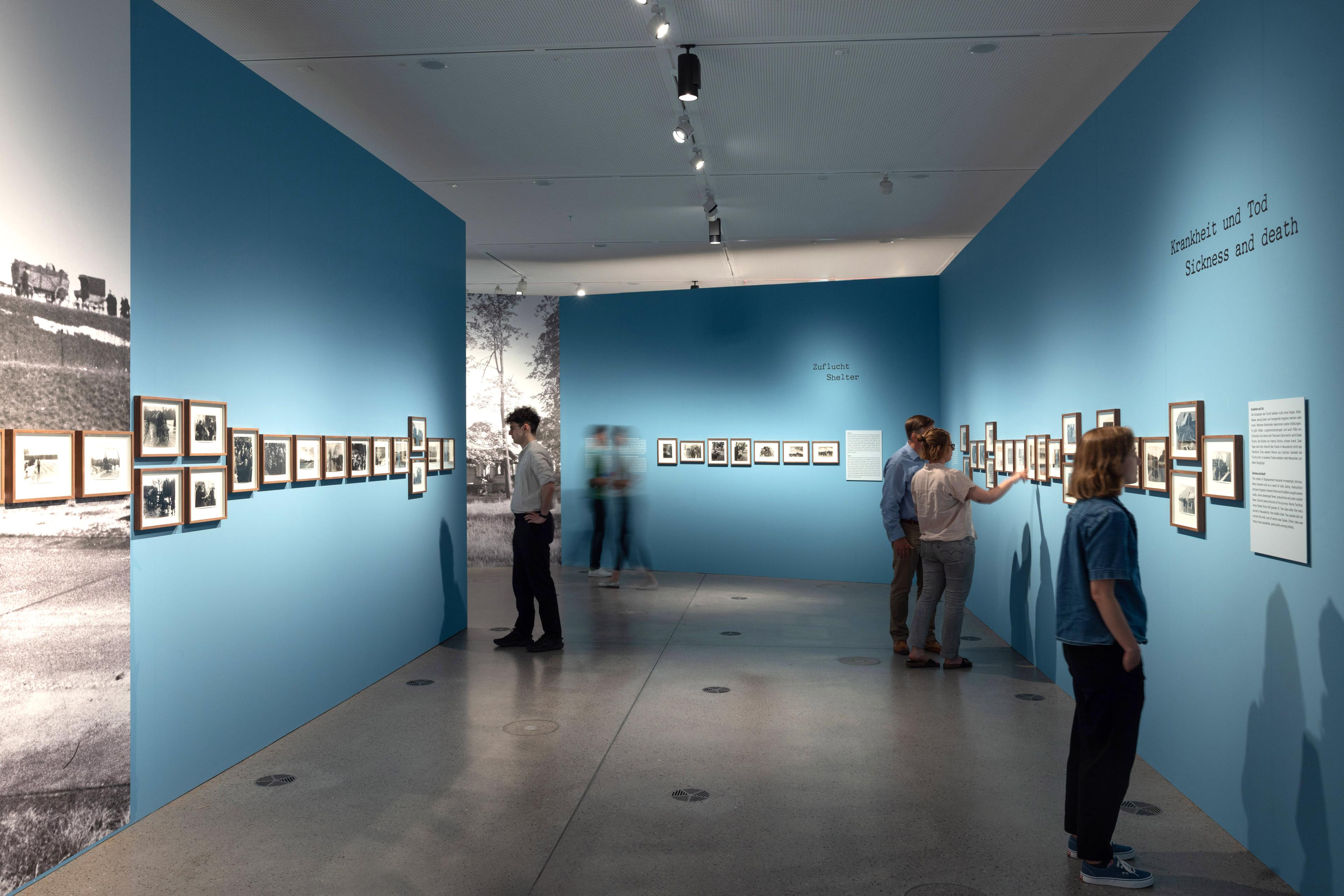 Ausstellungsansicht von "Der Treck". Mehrere Personen vor einer blauen Wand, an der eingerahmte Fotos hängen.