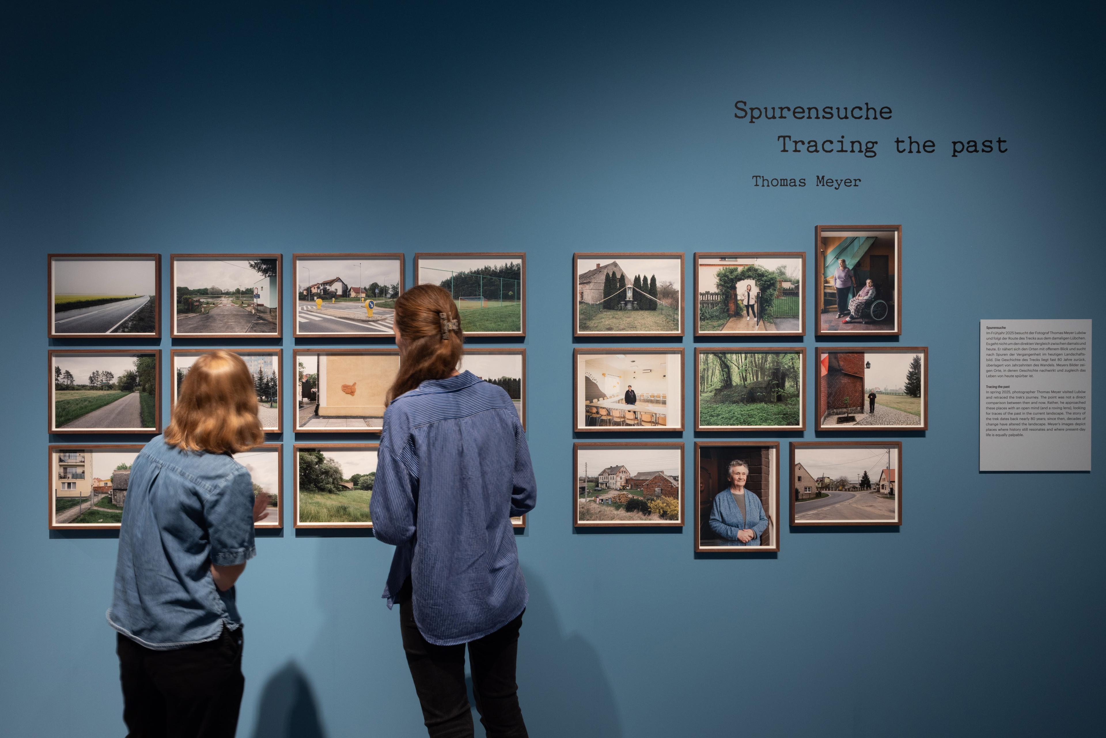 Ausstellungsansicht von "Der Treck". Zwei Frauen stehen vor einer blauen Wand, an der eingerahmte Fotos hängen.