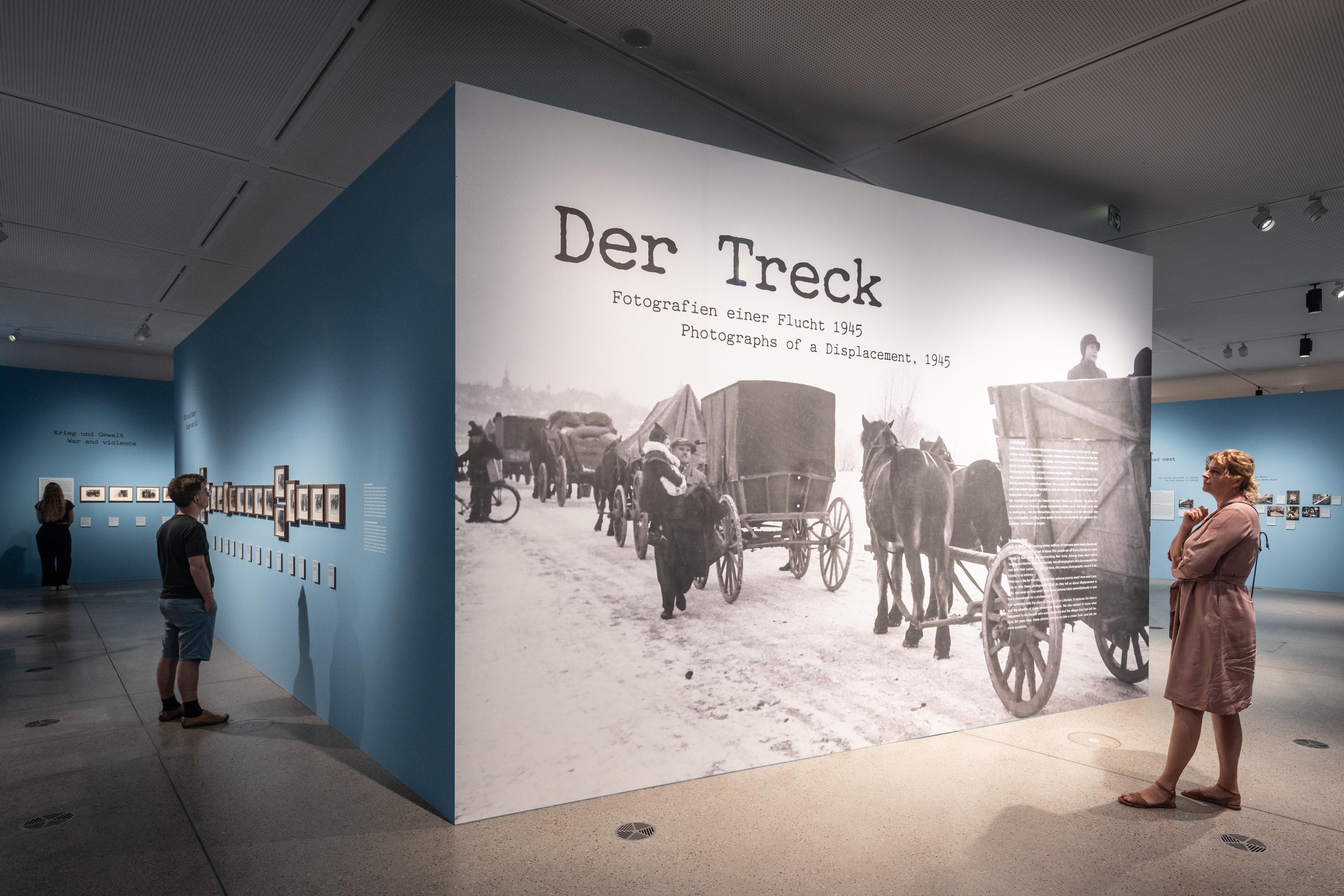 drei Personen, die die Ausstellung "Der Treck" anschauen, im Fokus ist eine große Ausstellungswand auf der der Titel groß zu sehen ist.