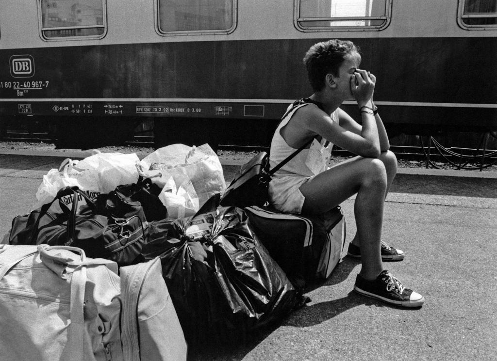 Schwarz-weiß Bild, ein Junge sitzt auf Gepäck an einem Bahngleis.