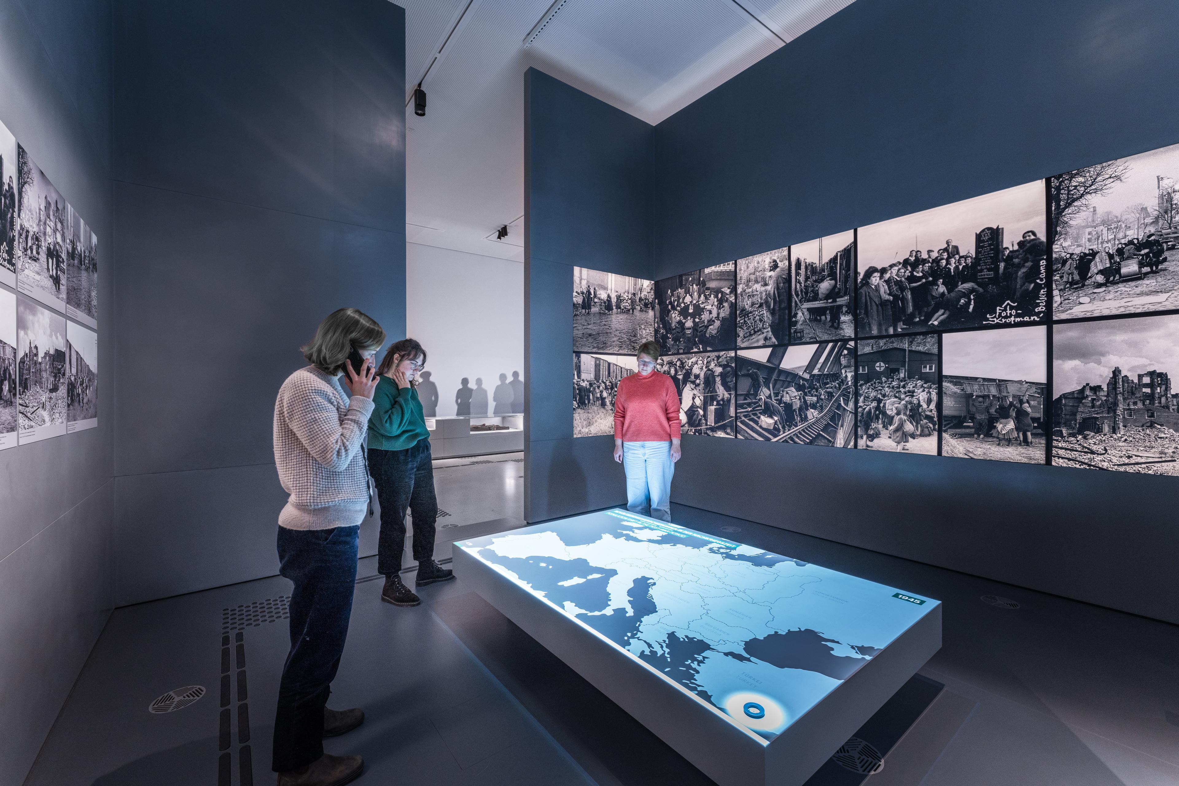 Drei Frauen stehen vor einer multimedial gestalteten Karte in einem Ausstellungsraum. An der Wand sind historische Fotos.