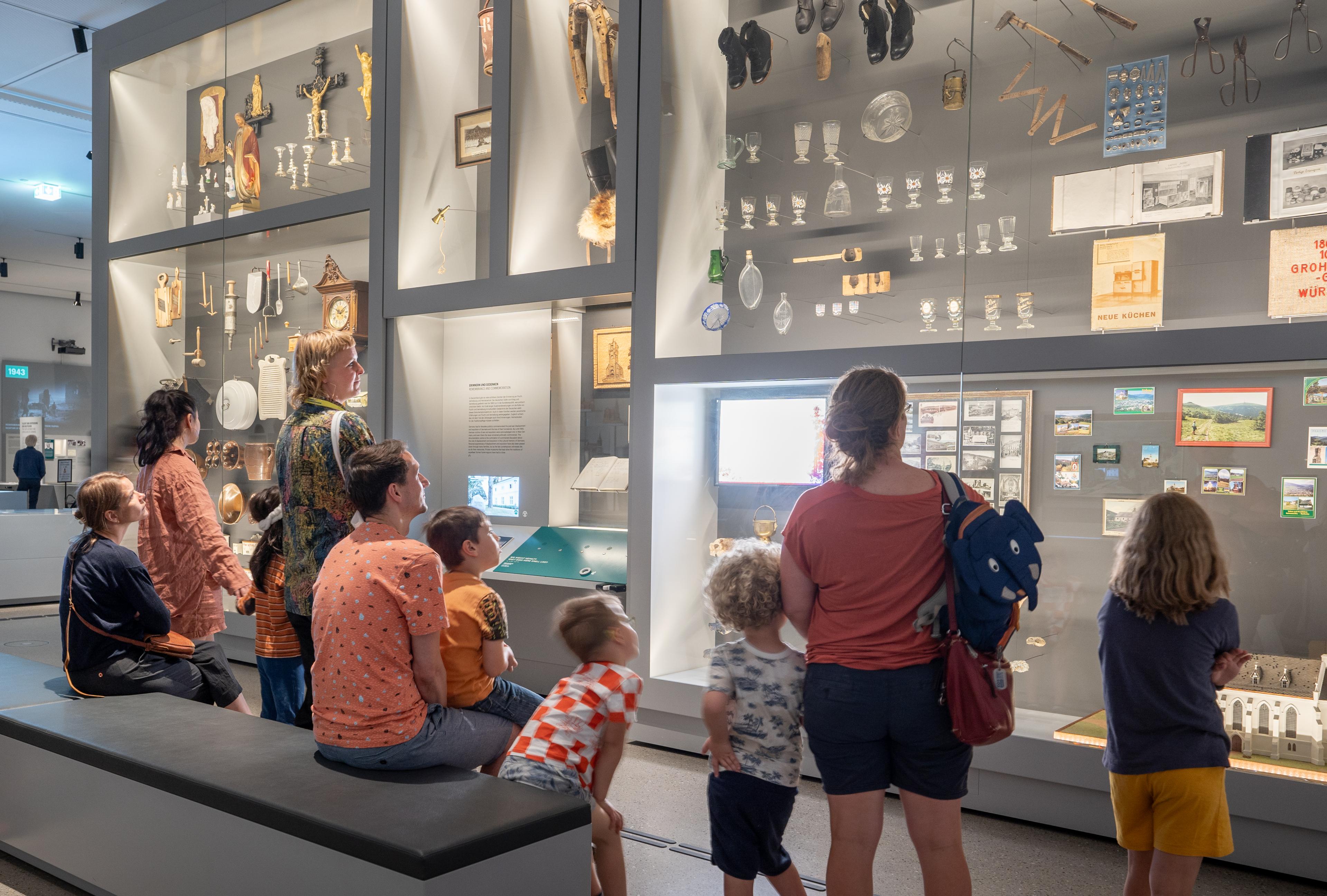 Eine Gruppe mit Kindern und Erwachsenen steht vor einer Ausstellungswand mit vielen Objekten.