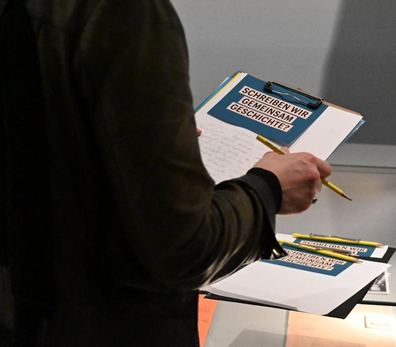 Nahaufnahme einer Person, die ein Klemmbrett in der Hand hält. darauf ist ein Blatt und eine Postkarte mit dem Schriftzug "Schreiben wir gemeinsam Geschichte?"