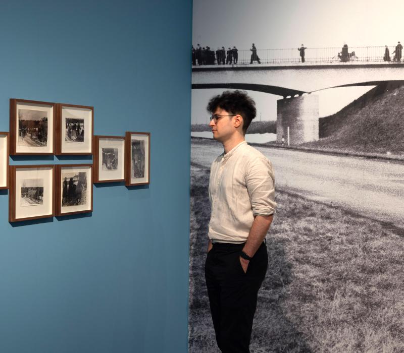 Ausstellungsansicht von "Der Treck". Ein Mann steht vor einer blauen Wand, an der eingerahmte Fotos hängen.