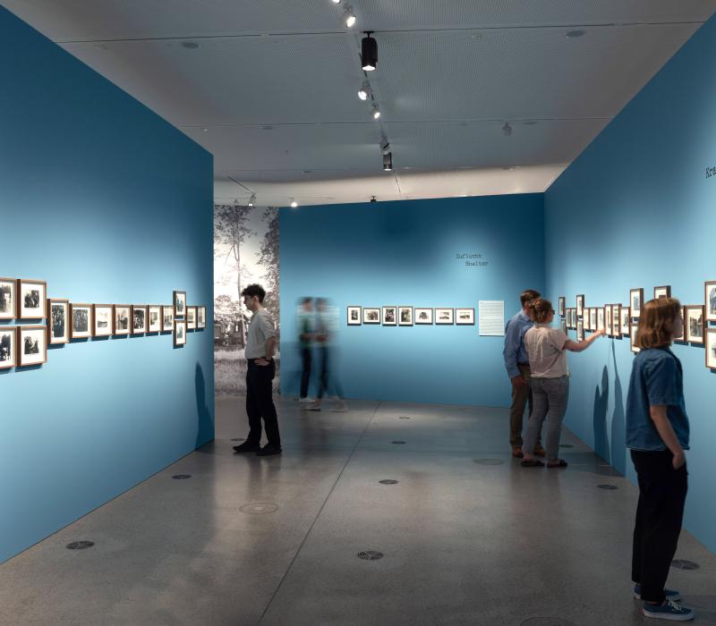 Ausstellungsansicht von "Der Treck". Mehrere Personen vor einer blauen Wand, an der eingerahmte Fotos hängen.