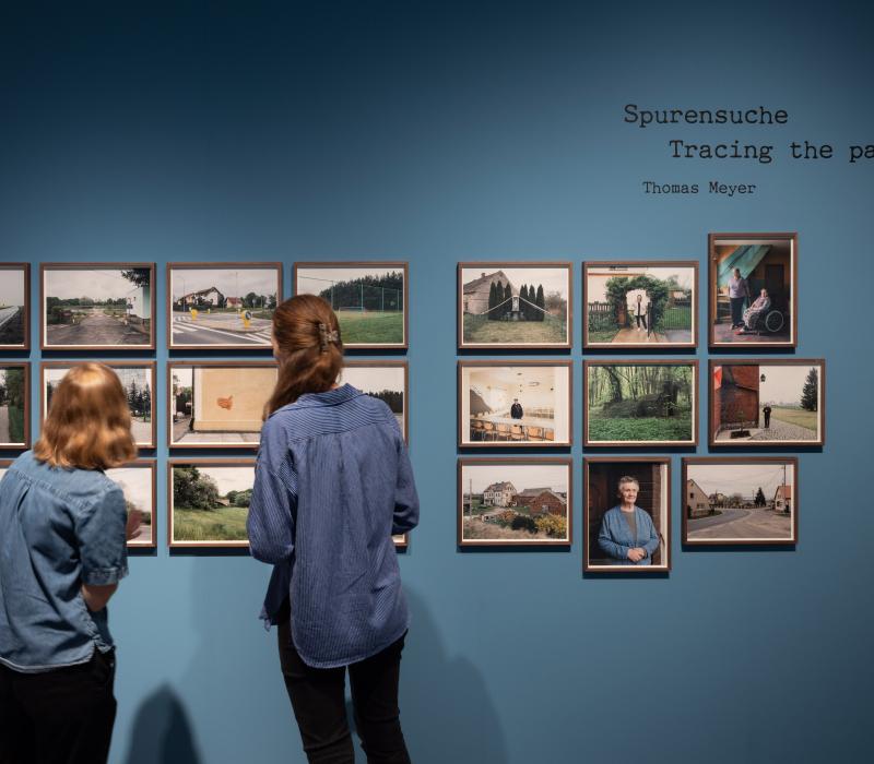 Ausstellungsansicht von "Der Treck". Zwei Frauen stehen vor einer blauen Wand, an der eingerahmte Fotos hängen.