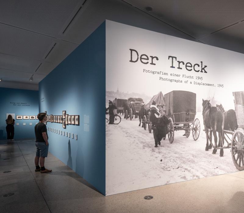 drei Personen, die die Ausstellung "Der Treck" anschauen, im Fokus ist eine große Ausstellungswand auf der der Titel groß zu sehen ist.