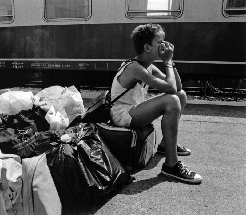 Schwarz-weiß Bild, ein Junge sitzt auf Gepäck an einem Bahngleis.