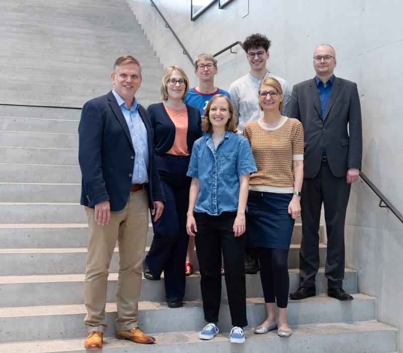Ein Gruppenbild von mehreren Personen (drei Männer, vier Frauen), die auf einer Treppe stehen und in die Kamera lächeln.