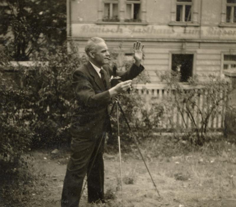 Schwarz-weiß Aufnahme eines Mannes, der mit Zigarre im Mund in einem Garten steht und eine Kamera auf einem Stativ bedient.