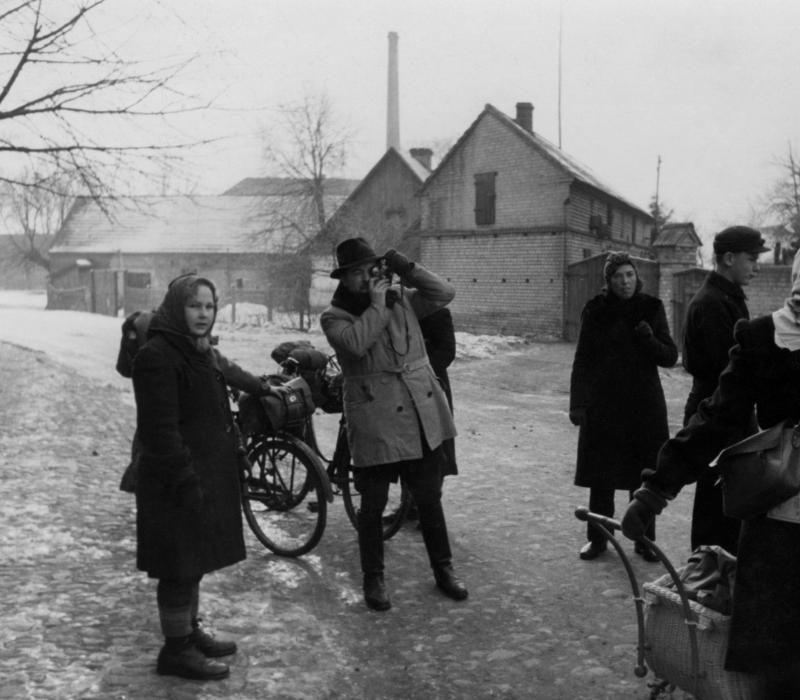 Schwarz-weiß Bild, zehn Personen, im Hintergrund sind einige Häuser. Zwei Personen sind im Fokus, ein Mann hält eine Kamera, eine Frau hält mit einer Hand ein Fahrrad fest