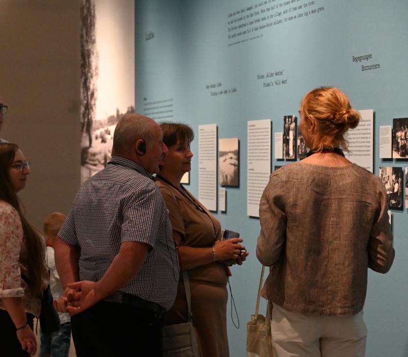 Eine Frau steht vor mehreren Bildern einer Ausstellung und erzählt einer Gruppe darüber.