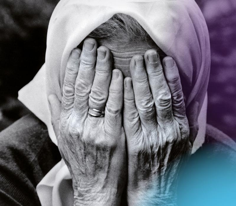 Schwarz-weiß Porträt einer älteren Frau mit weißem Kopftuch, die die Hände vors Gesicht hält.