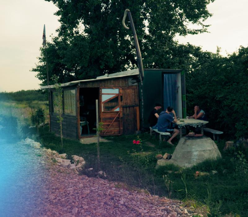 Drei Männer sitzen am Tisch im Gespräch vor einer kleinen Hütte an einer Straße.
