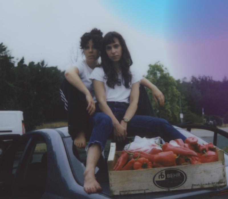 zwei Frauen sitzen auf einem Auto, vor ihnen liegt eine offene Kiste mit roter Paprika.