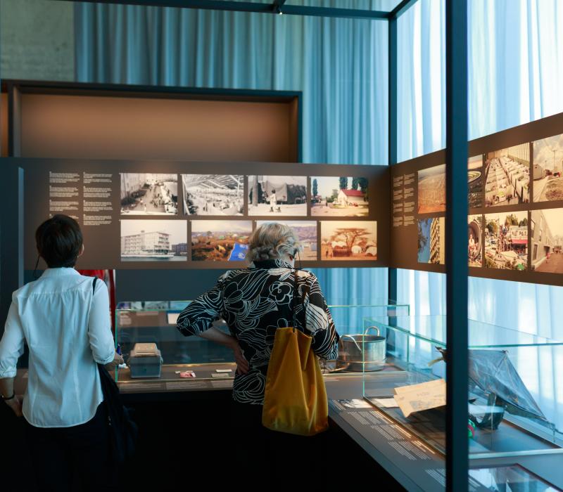 Rückenansicht von zwei Personen, die gerade vor einer Ausstellungswand stehen und Bilder anschauen.