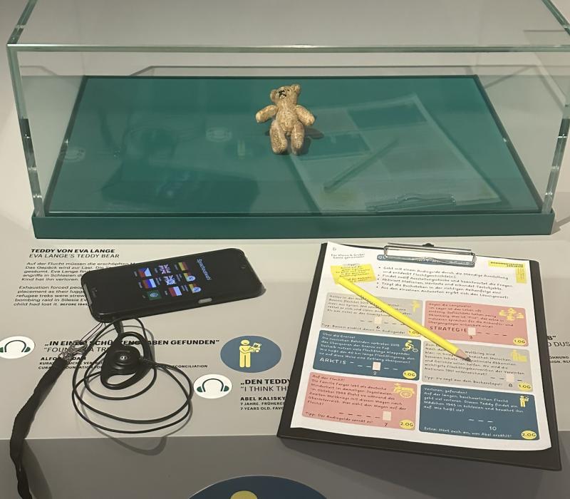 Auf einer Museumsvitrine in der ein Teddy ist, liegt ein Audio-Guide und ein Papier mit Quiz-Fragen und einem Bleistift.