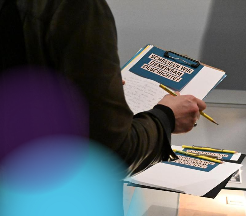 Eine Person hält einen Block und Stift in der Hand, man sieht die Person von Hinten, der Fokus ist auf dem Papier. Darauf ist der Text "Schreiben wir gemeinsam Geschichte?" zu sehen.