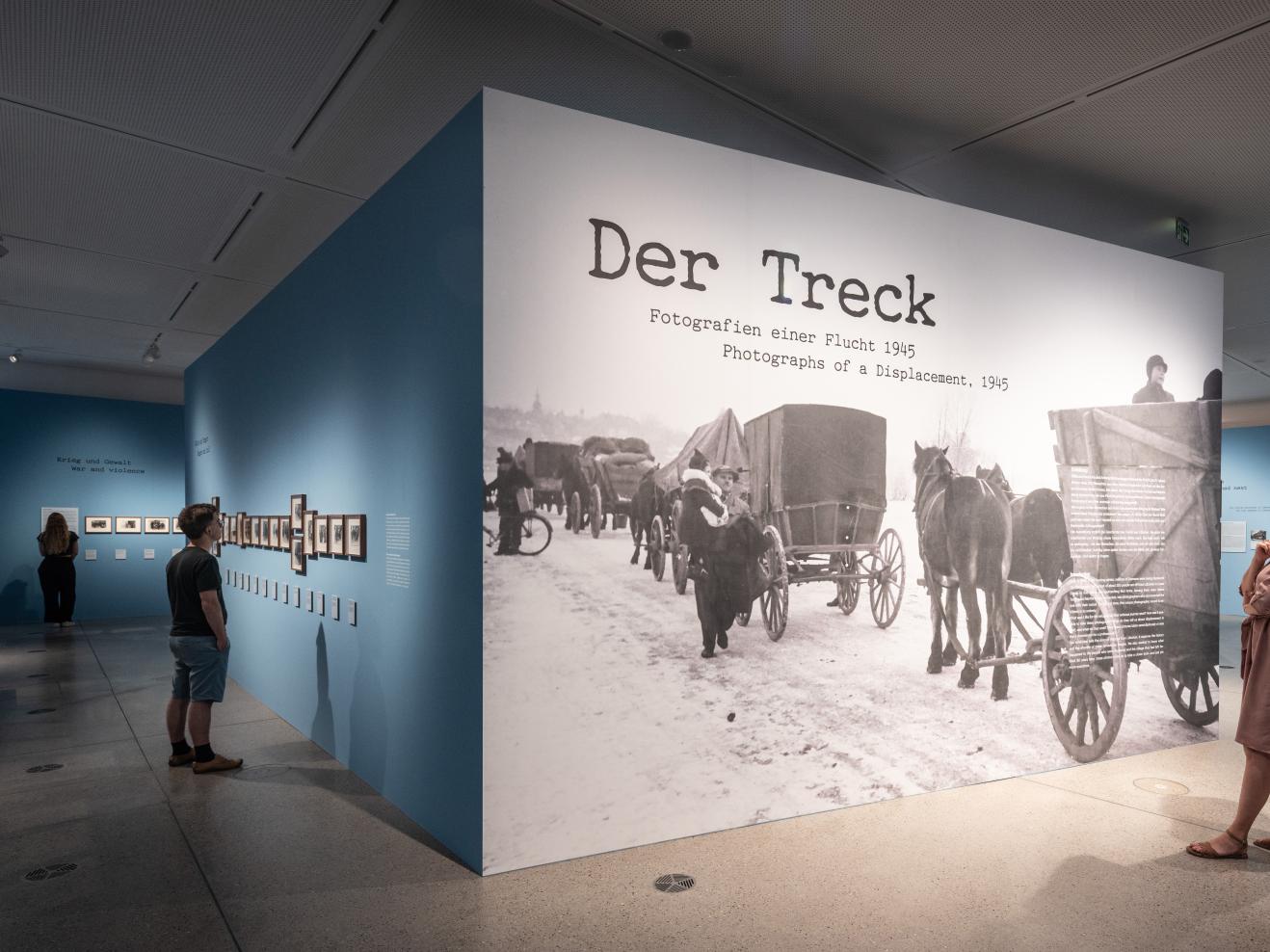 drei Personen, die die Ausstellung "Der Treck" anschauen, im Fokus ist eine große Ausstellungswand auf der der Titel groß zu sehen ist.